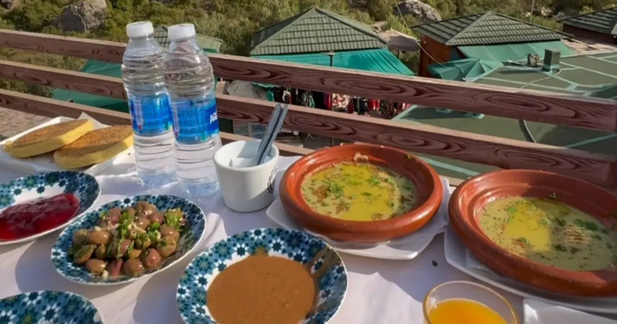 Petit-déjeuner marocain dressé sur une table, devant la rambarde en bois d'une terrasse et les toits verts d'Akchour